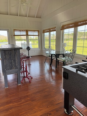 A room with wooden floors, a table with chairs, a fan, and a window with a view of greenery.