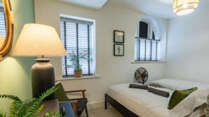 Quarto com cama, escrivaninha, luminária, ventoinha e duas janelas com persianas.