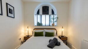 Quarto com cama, duas luminárias de cabeceira, janela com persianas e uma planta no parapeito.