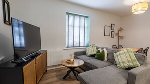 Uma sala de estar moderna com uma TV de tela plana, um sofá cinza, uma pequena mesa de centro de madeira e uma janela com persianas.