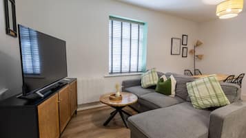 A modern living room with a flat-screen TV, a grey sofa, a small wooden coffee table, and a window with blinds.
