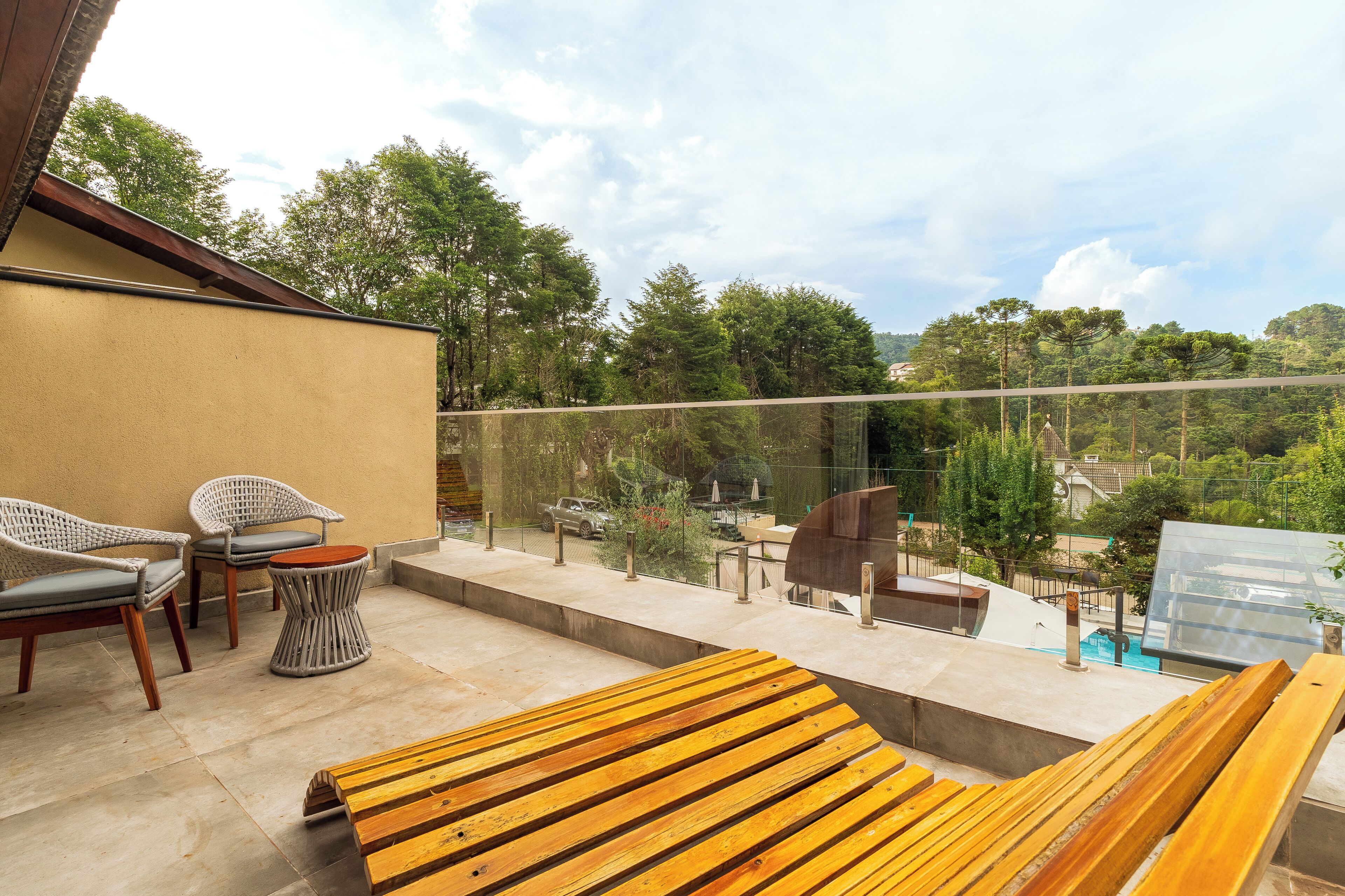 A balcony with wooden furniture, a glass railing, and a view of trees and buildings.