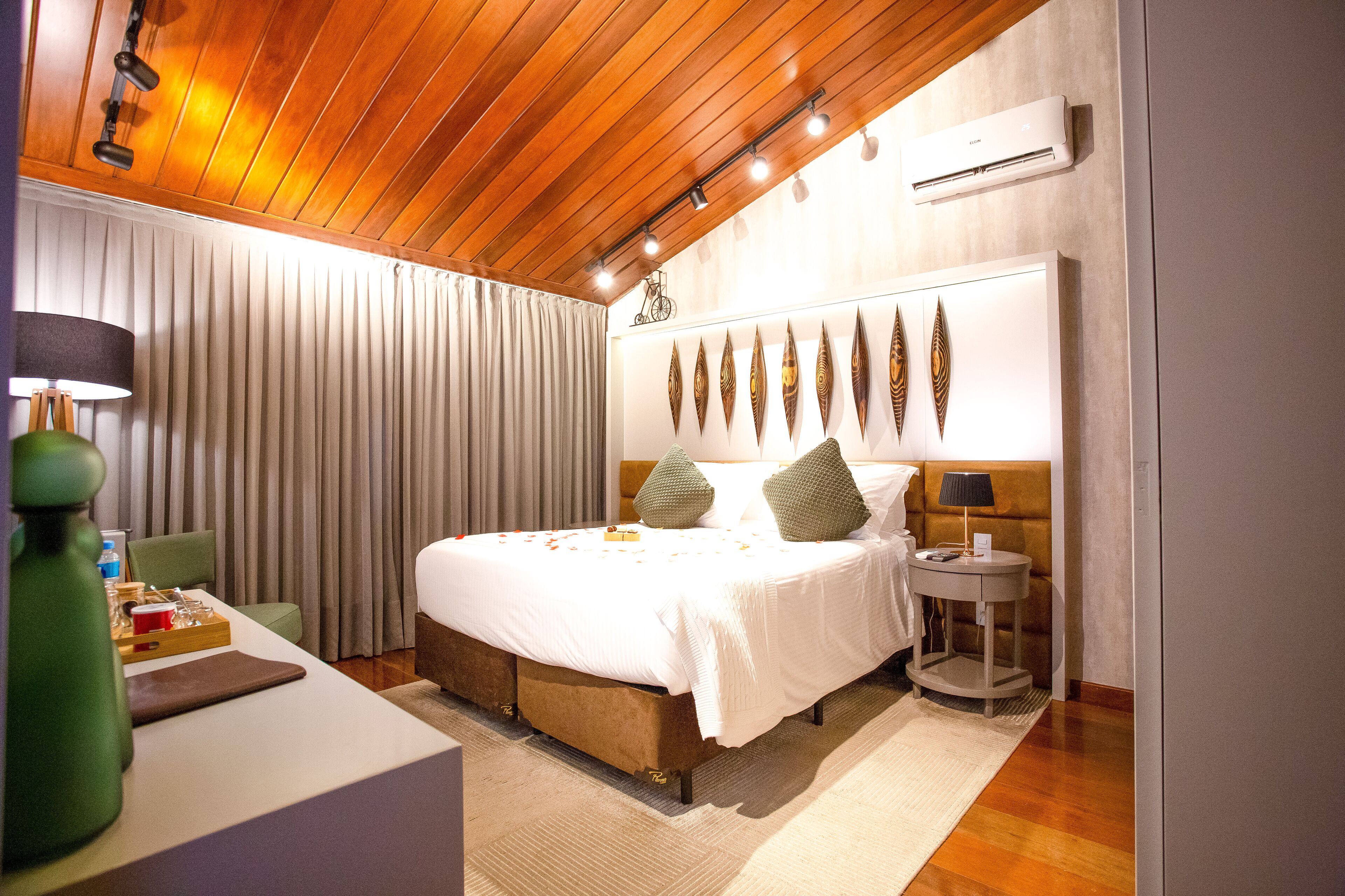 A hotel room with a wooden ceiling, a bed with white bedding, a bedside table with a lamp, and a wall-mounted air conditioner.