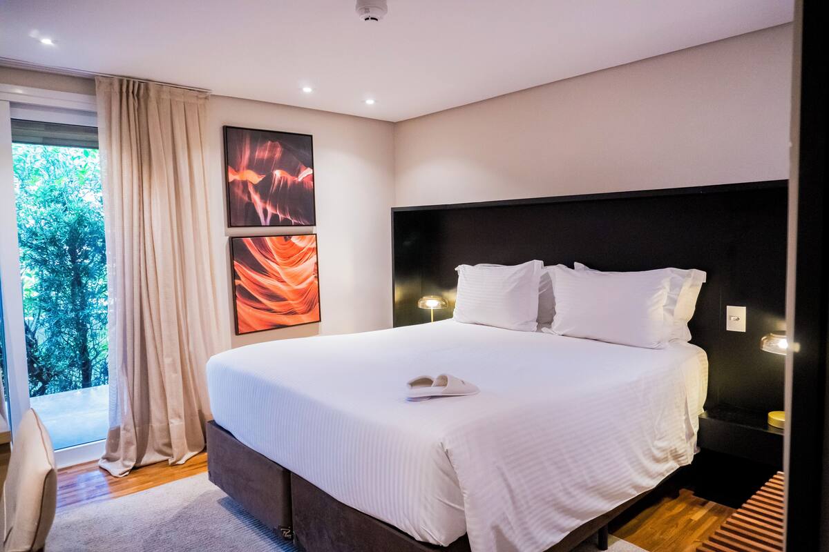 Una camera d'albergo moderna con un letto grande, vista sugli alberi dalla finestra e opere d'arte alle pareti.