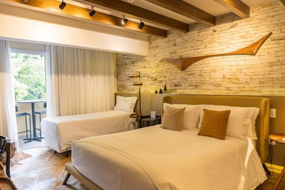 Camera d'albergo con un letto grande, un letto più piccolo, una testiera in legno, una parete in mattoni e una finestra con tende.