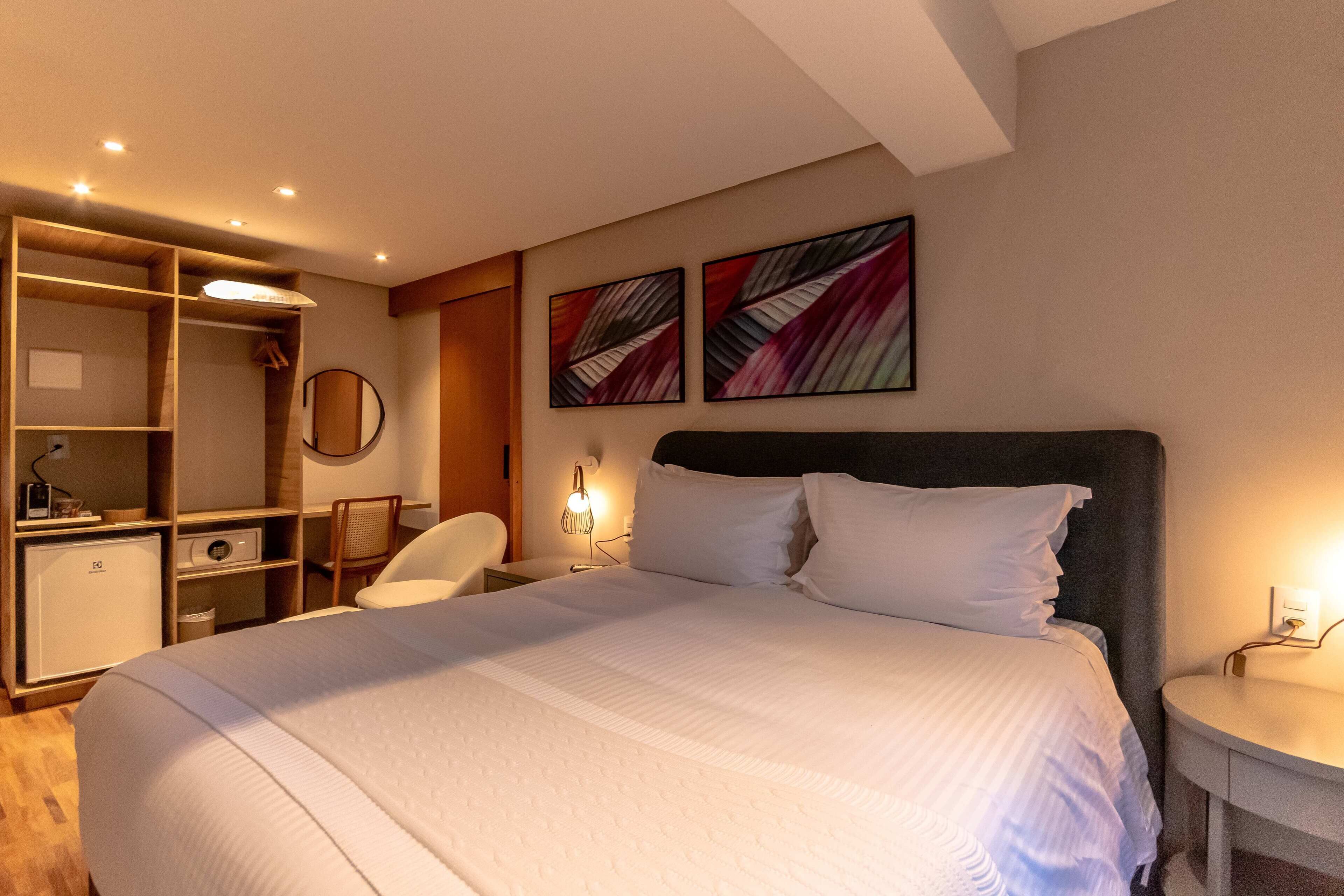 A hotel room with a large bed, a desk, a chair, and two abstract paintings on the wall.