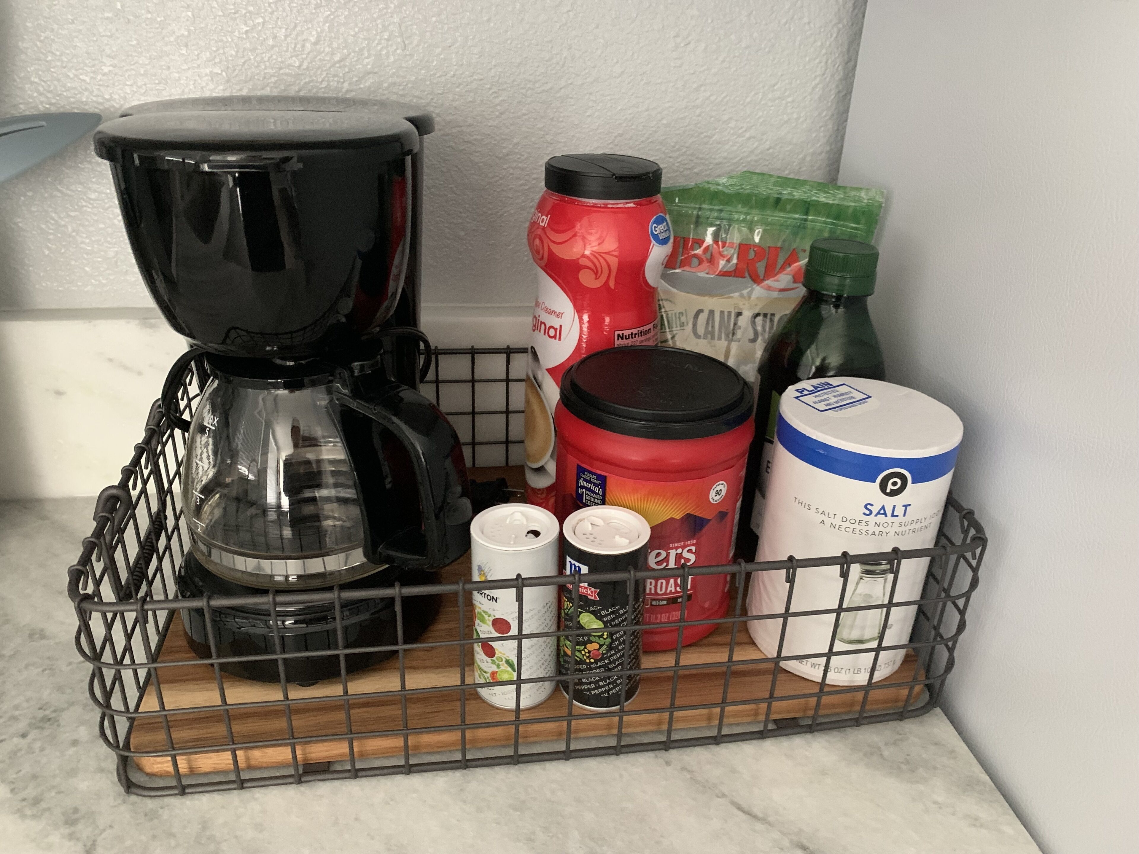 A kitchen counter with a coffee maker, a salt container, a can of black pepper, a container of sugar, a bag of cane sugar, and a bag of flour.