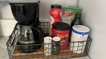 A kitchen counter with a coffee maker, a salt container, a can of black pepper, a container of sugar, a bag of cane sugar, and a bag of flour.