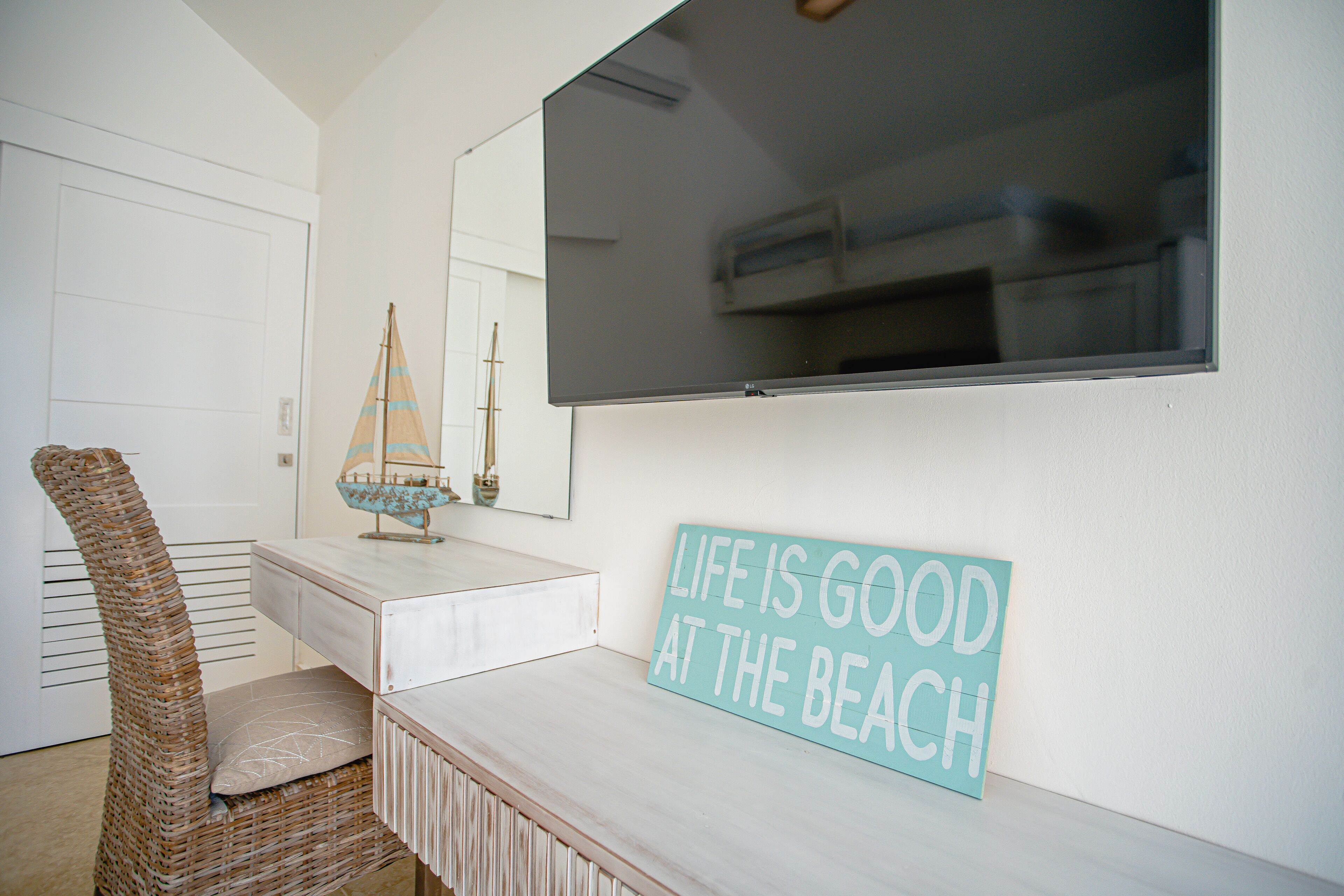 A room with a white wall, a mounted TV, a wicker chair, a small ship model, and a sign that reads "LIFE IS GOOD AT THE BEACH".