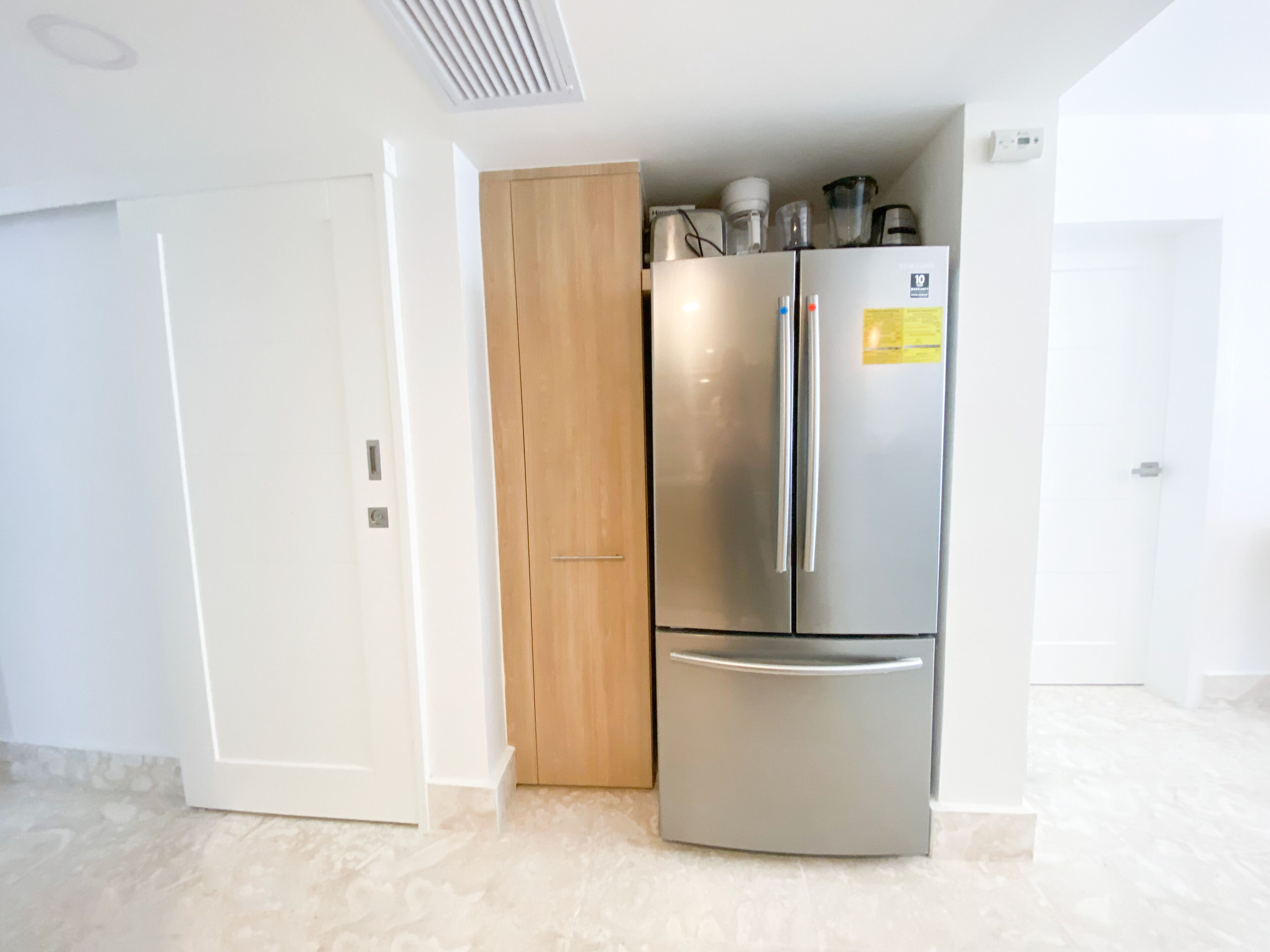 A compact kitchen with a built-in stainless steel refrigerator and oven, a wooden door, and a white tiled floor.