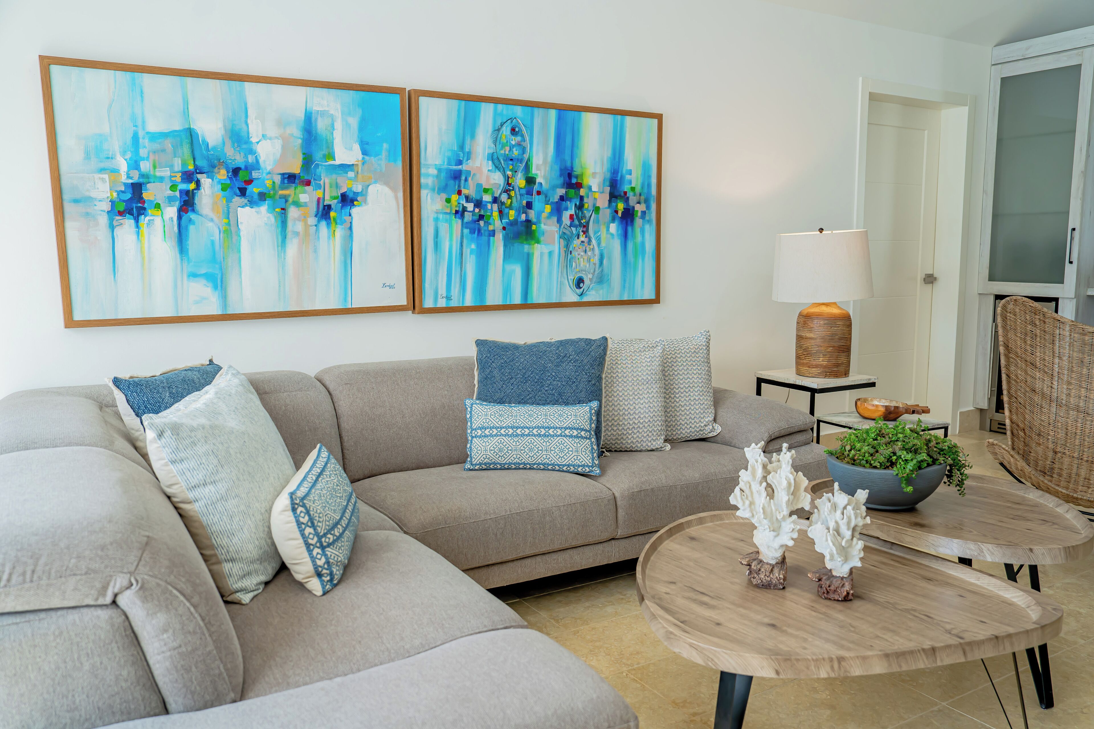A living room with a sofa, two abstract paintings, a lamp, a small table with decorative items, and a wicker chair.