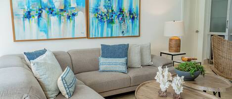 A living room with a sofa, two abstract paintings, a lamp, a small table with decorative items, and a wicker chair.