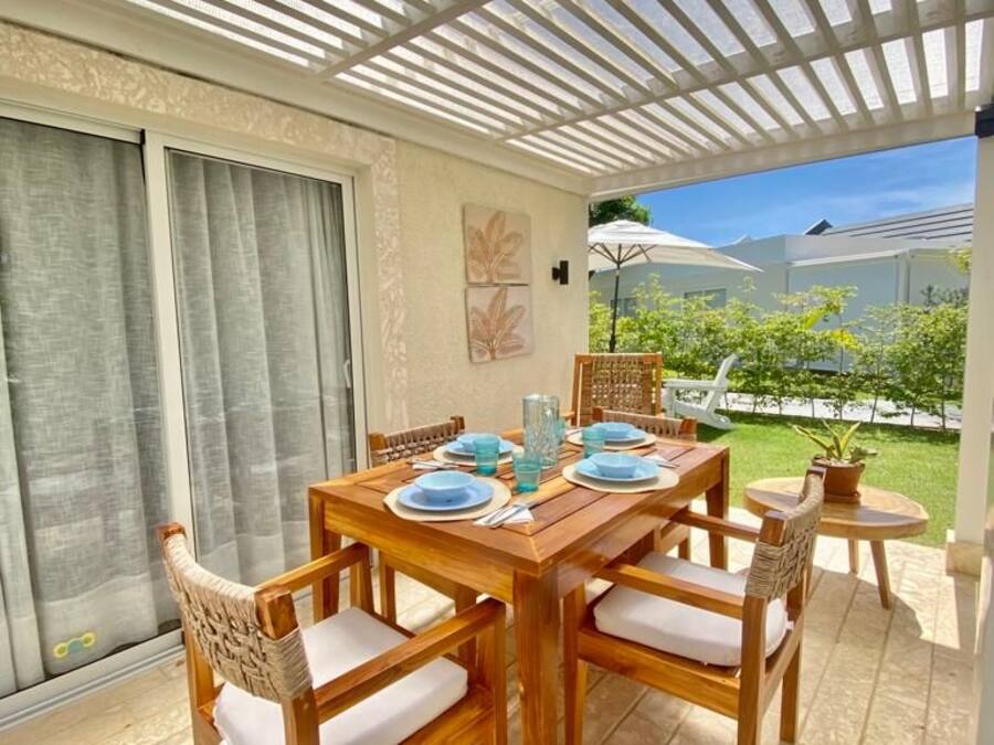 An outdoor dining area with a wooden table and chairs, a pergola overhead, and a view of a garden.