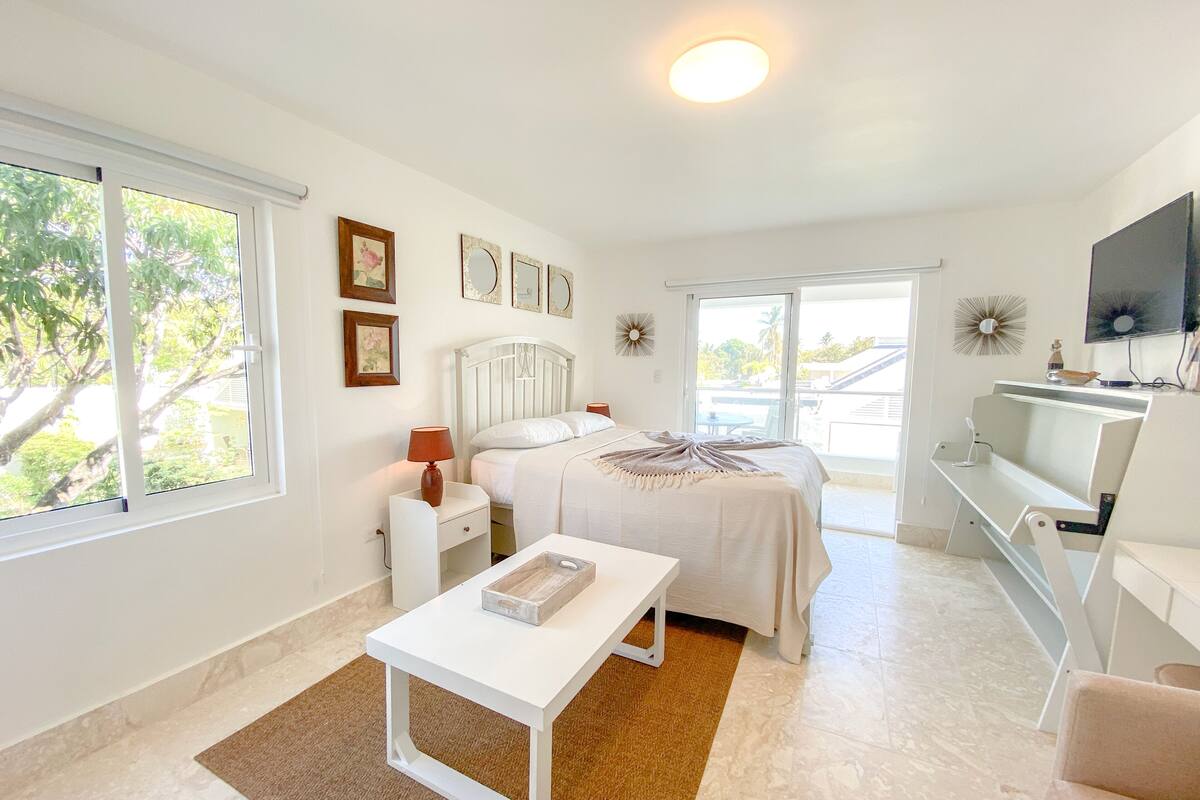 Une chambre à coucher comprenant un lit, des tables de chevet, un bureau, une télévision et une fenêtre donnant sur des arbres.