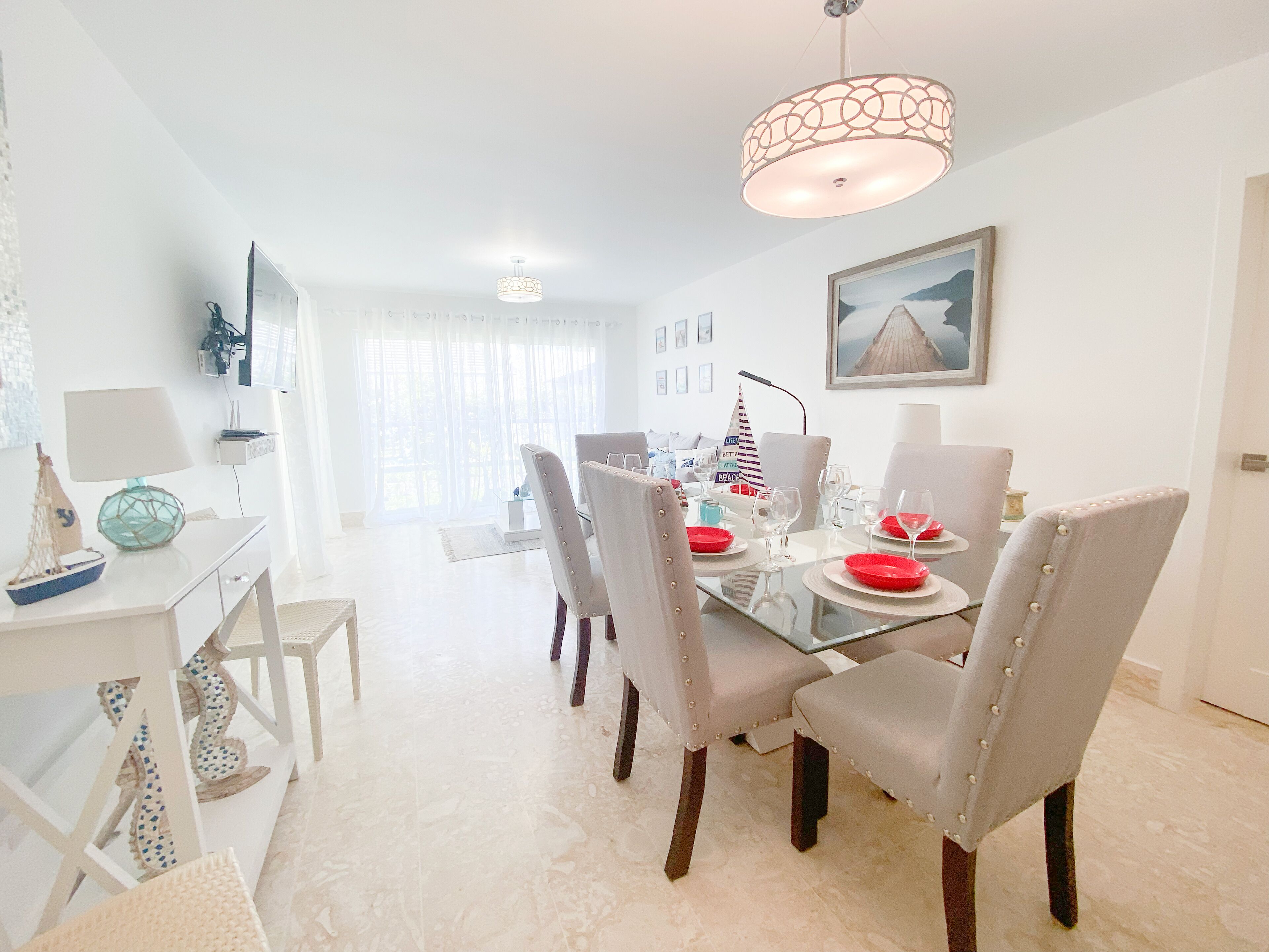 A dining area with a glass table, white chairs, and a chandelier. There are decorative items on the table and a view of the living room in the background.