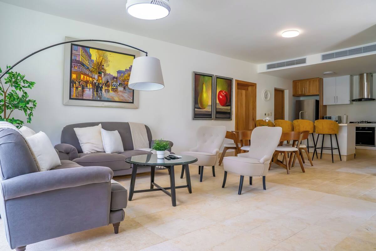 Un salon moderne avec un canapé, des fauteuils, une table basse et un coin repas avec des chaises. Un tableau orne le mur et on aperçoit une cuisine au fond.