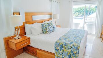 Ein Schlafzimmer mit einem hölzernen Kopfende, einem Bett mit weißer Bettwäsche und einem blumigen Tagesdecken in Blau und Gelb, zwei Nachttischlampen und Blick auf einen Balkon mit weißen Korbmöbeln.