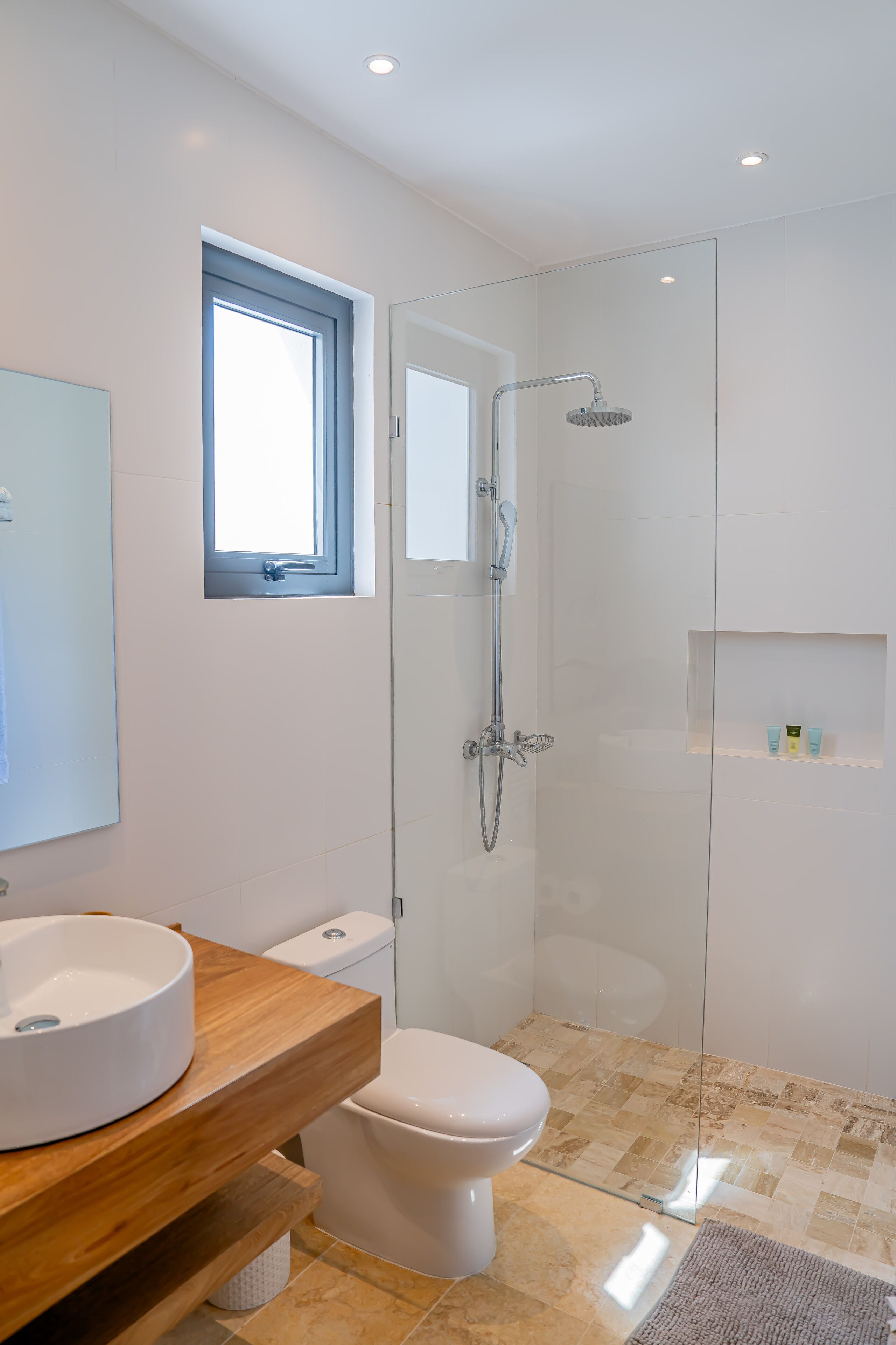 A modern bathroom with a glass-enclosed shower, a toilet, a sink with a wooden vanity, and a small window.