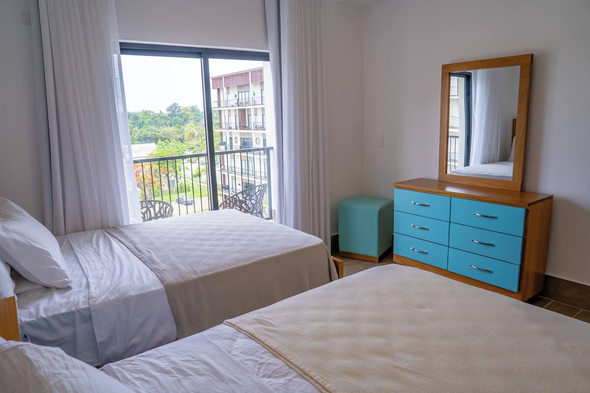 Une chambre d’hôtel avec deux lits, une fenêtre avec des rideaux, une commode en bois avec des tiroirs et un balcon avec une balustrade.