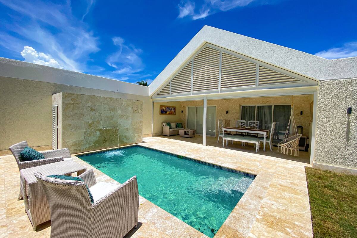 Un espace piscine doté d’une piscine à l’eau cristalline, de fauteuils de jardin en osier et d’un patio couvert avec une table et des chaises.