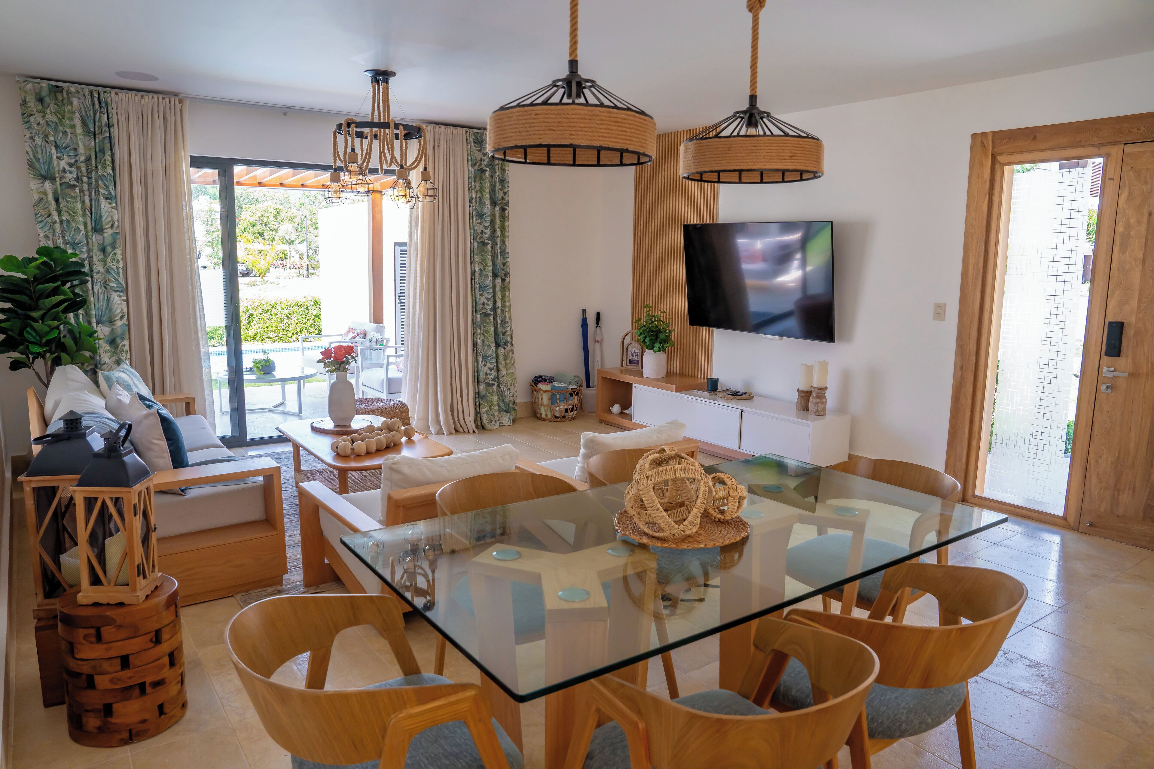 A modern living room with a glass-top dining table, wooden chairs, a sofa, and a TV mounted on the wall.