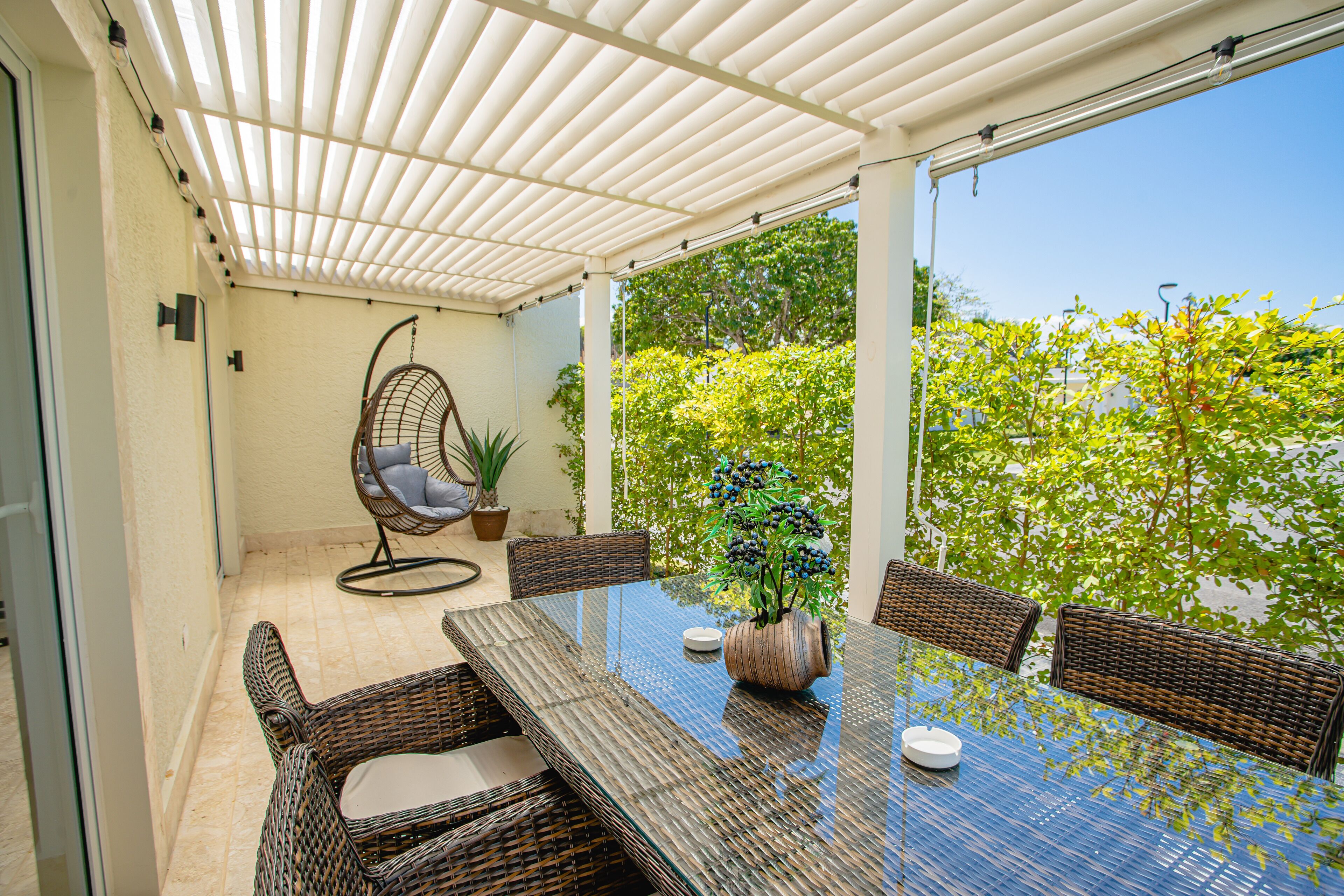 Un patio cubierto con una mesa de vidrio y sillas de mimbre, rodeado de follaje y un cielo despejado.