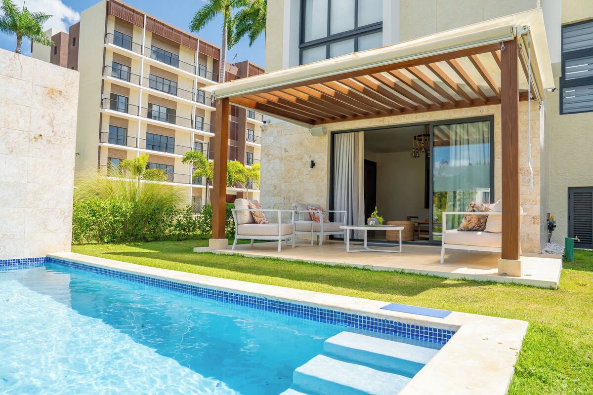Un espace aménagé au bord de la piscine, avec un coin salon couvert, une piscine et un bâtiment doté de balcons en arrière-plan.