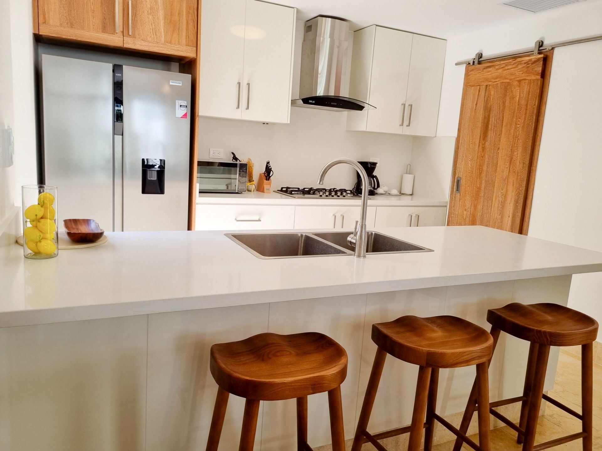 Una cocina moderna con refrigerador de acero inoxidable, armarios blancos, pileta doble y taburetes de madera.