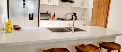 Una cocina moderna con refrigerador de acero inoxidable, armarios blancos, pileta doble y taburetes de madera.