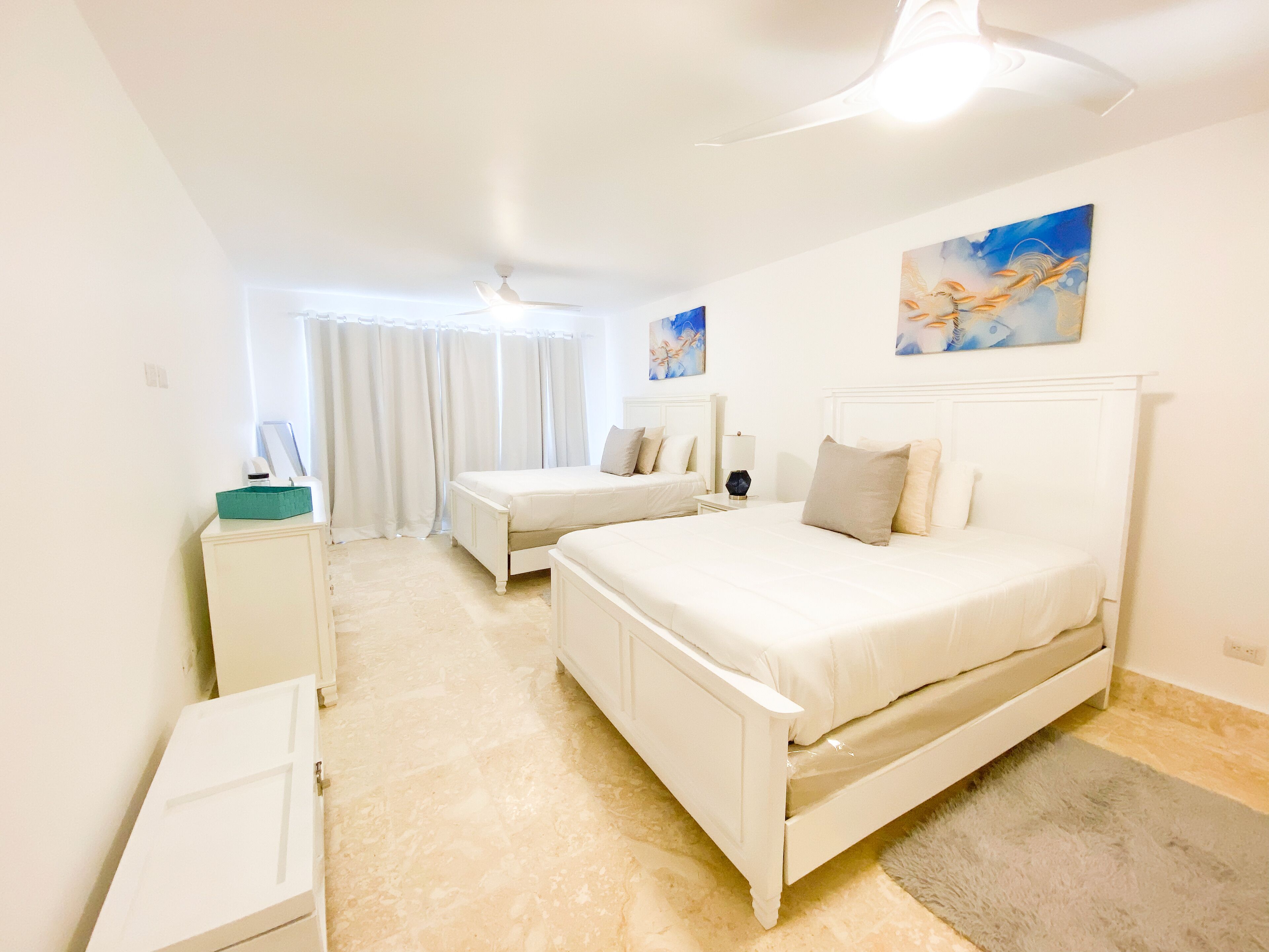A bedroom with two beds, a bedside table, a nightstand, a ceiling fan, and artwork on the wall.
