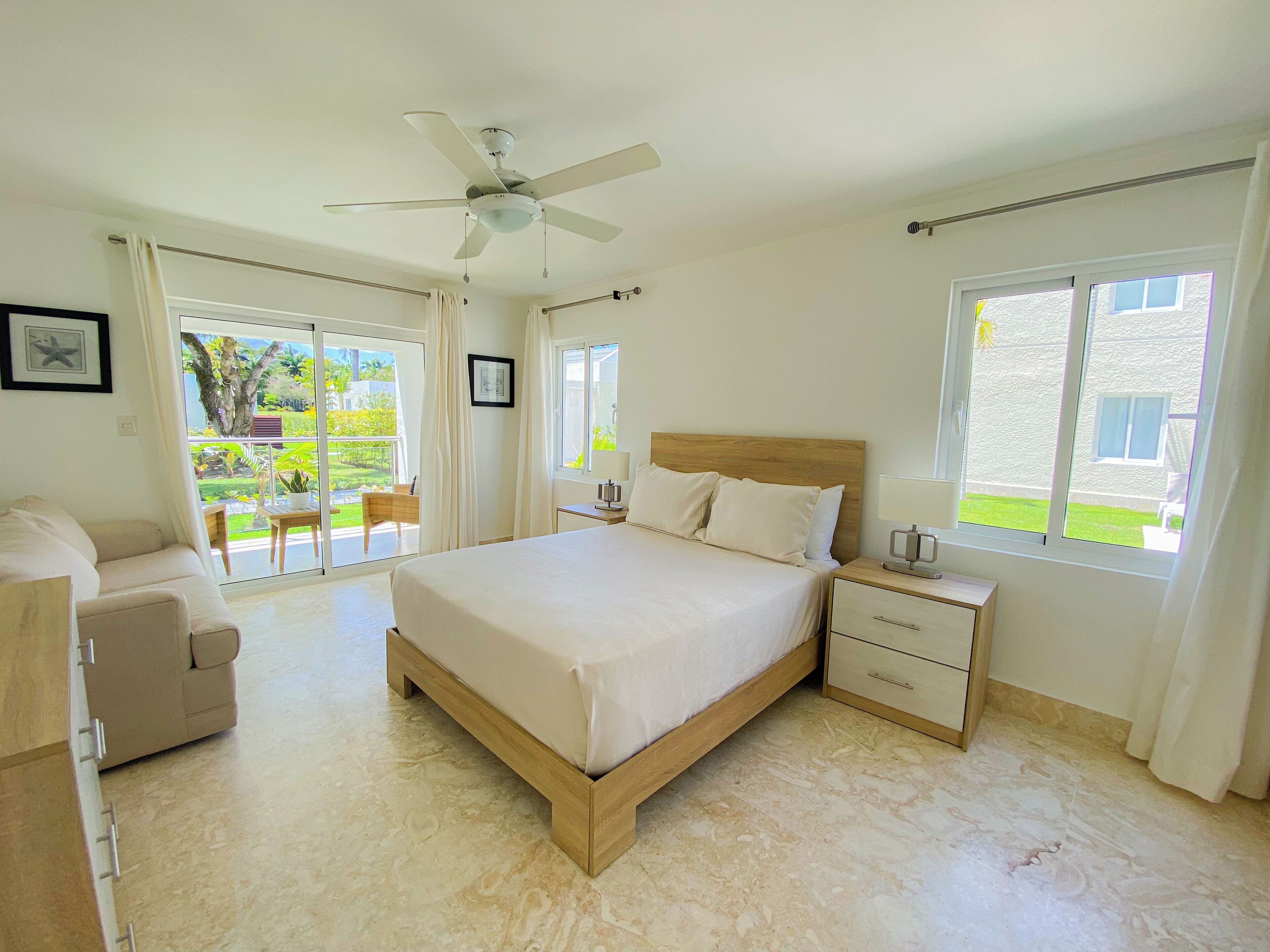 A spacious bedroom with a large bed, a ceiling fan, and a view of a garden through the sliding glass door.
