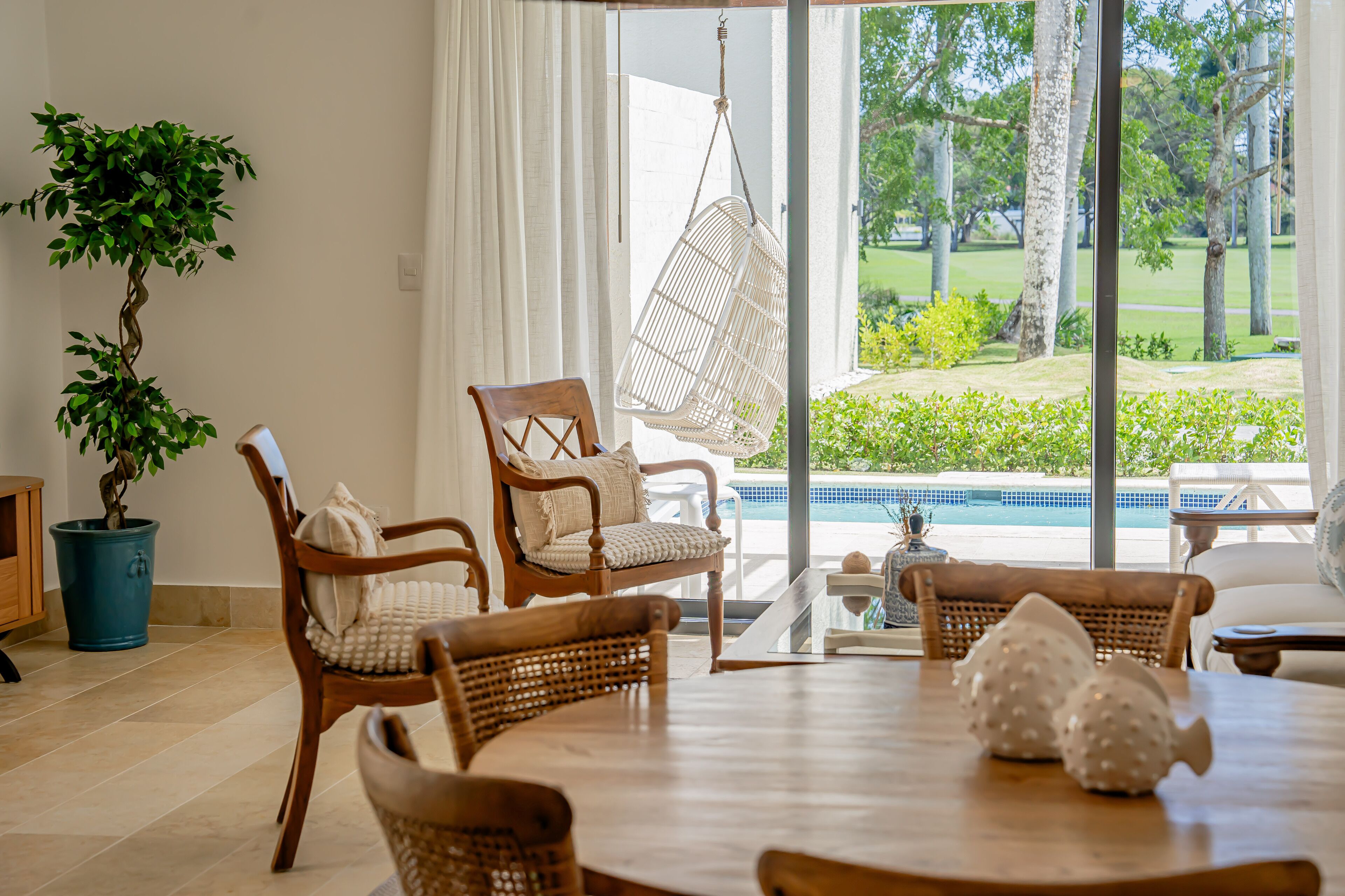Un salon spacieux doté d’une grande fenêtre donnant sur une piscine et un jardin. On y trouve plusieurs chaises en bois et une table à manger agrémentée de sculptures de poissons décoratives.