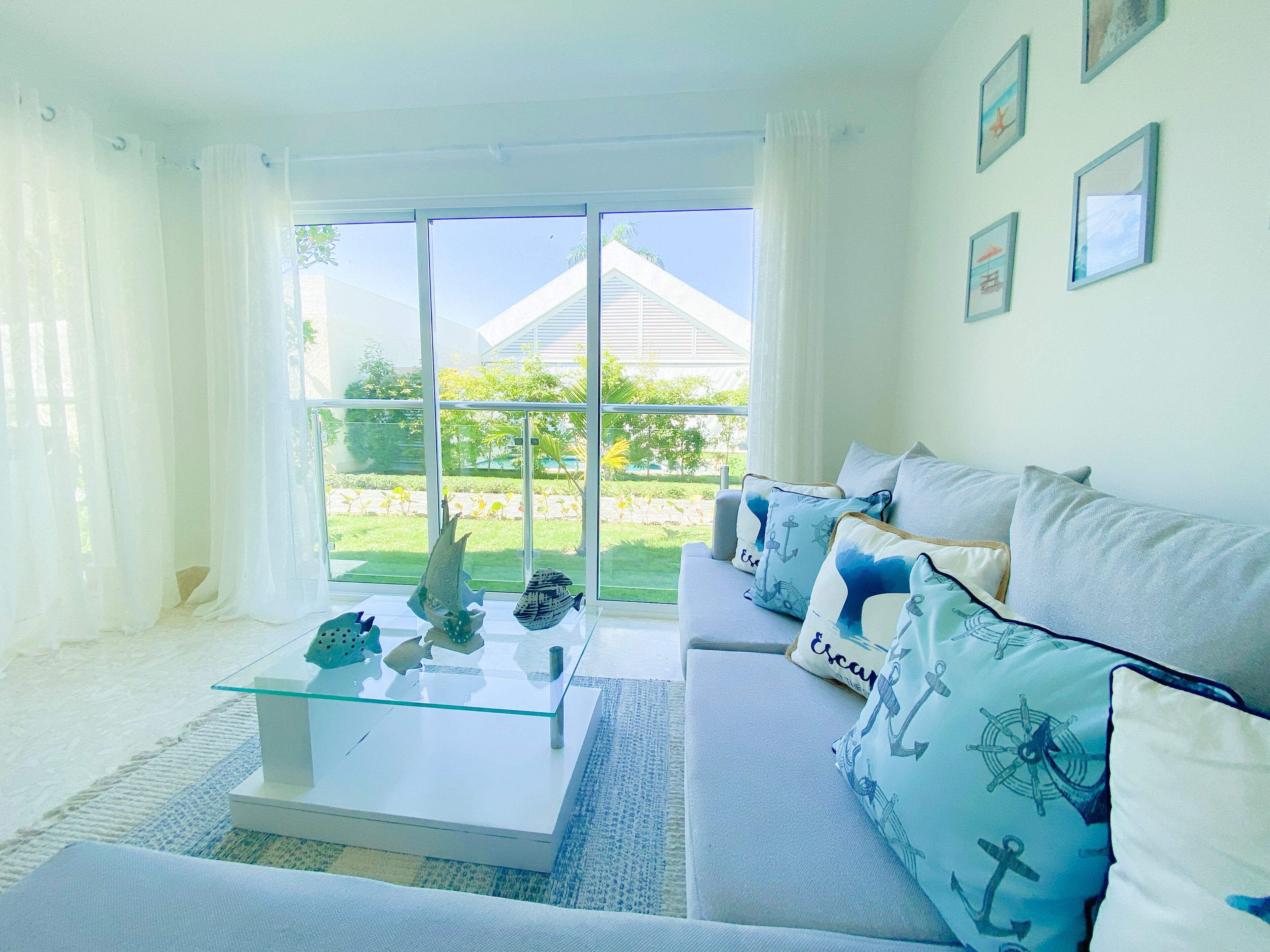A bright living room with a glass coffee table, a white sofa with nautical-themed cushions, and a view of a house and greenery outside.