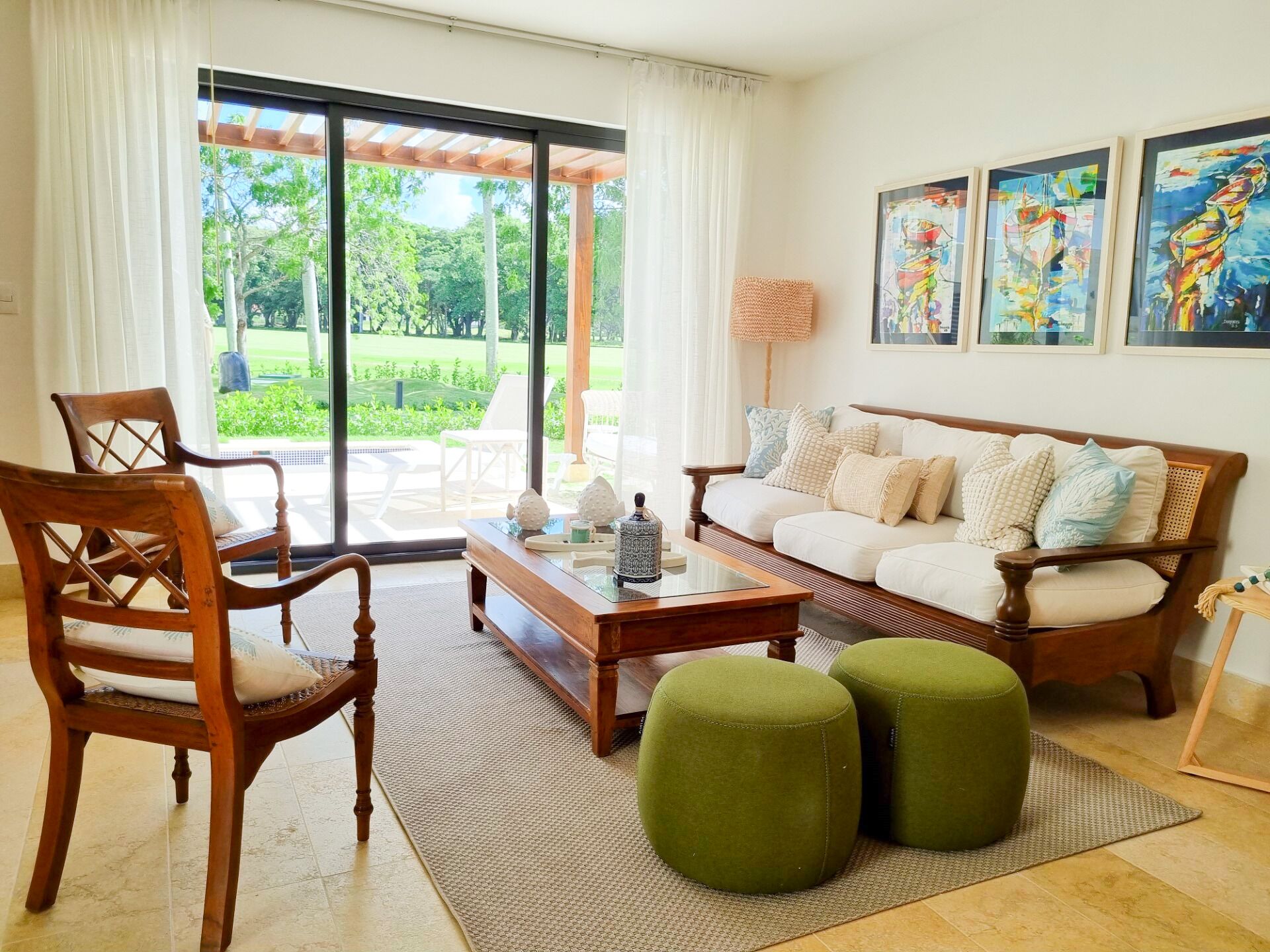 A living room with a sofa, two ottomans, a wooden coffee table, and a chair. There is a large window with a view of greenery outside.
