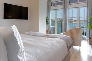 Ein Hotelzimmer mit einem großen Bett, einem an der Wand befestigten Fernseher, Meerblick durch große Fenster und einem gestreiften Sessel.