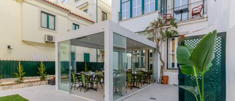 A modern outdoor dining area with glass walls, surrounded by a well-maintained garden and a white building with green accents.