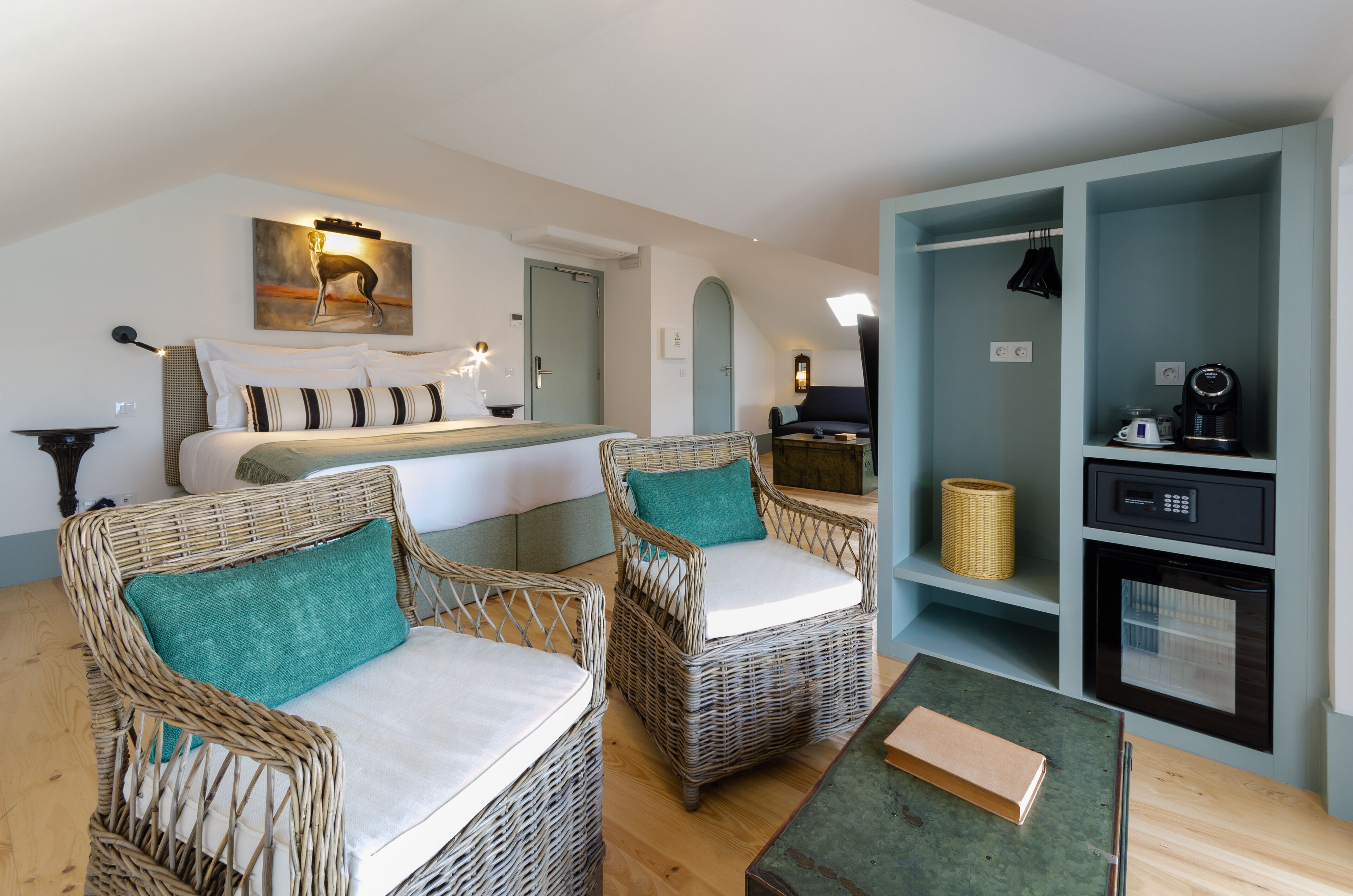 A modern hotel room with a large bed, two wicker chairs with teal cushions, a small table, and a kitchenette area.