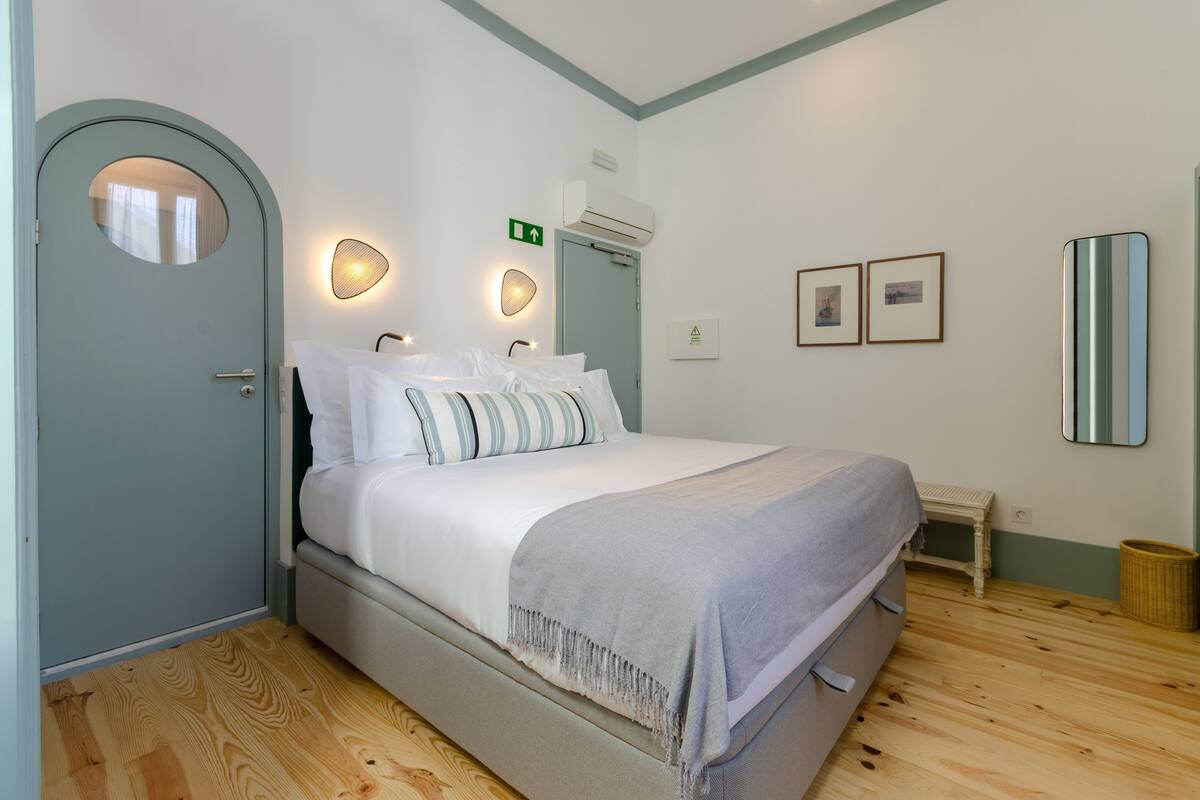 A bedroom with a bed, a door, an air conditioning unit, a small table, and framed pictures on the wall.