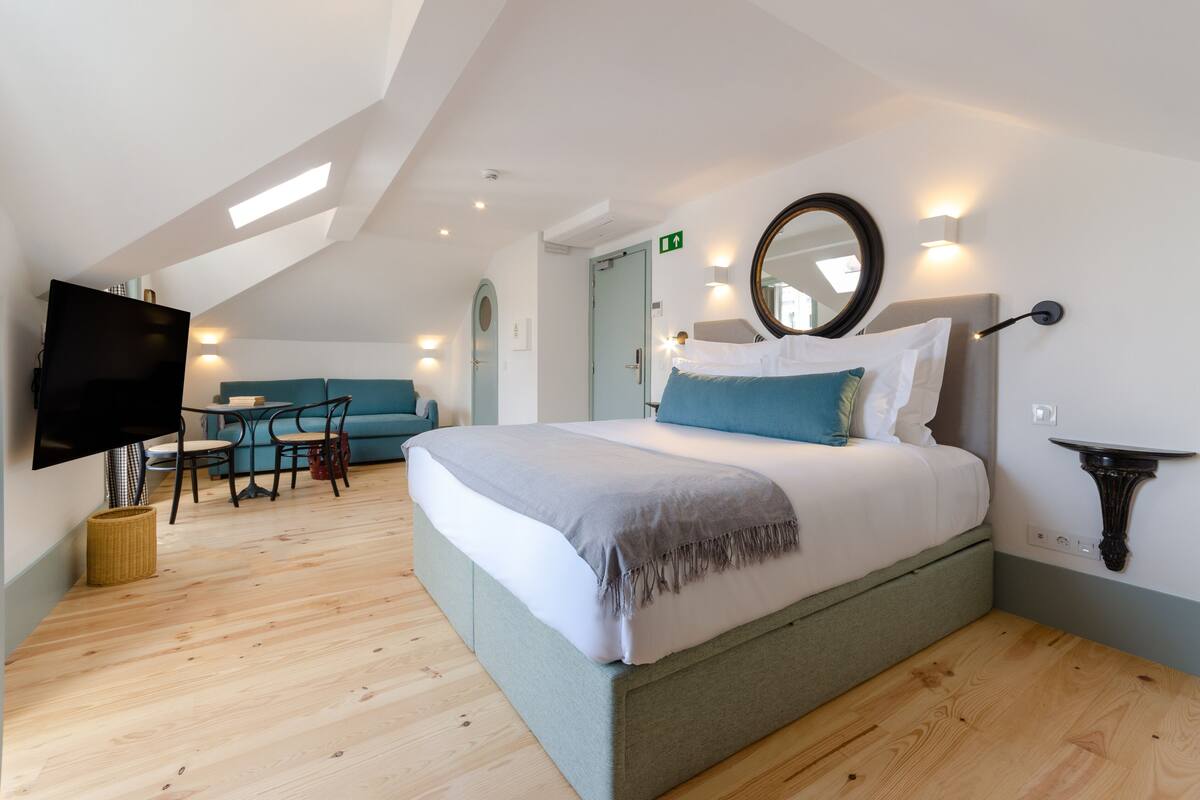 A modern hotel room with a large bed, a flat-screen TV, a dining area with a table and chairs, and a ceiling with skylights.