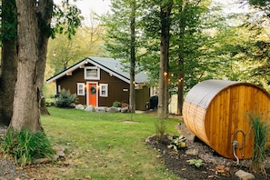 Een rustieke hut met een houten gevel, een rode deur en een grote houten vat in de tuin.
