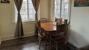 Une salle à manger avec une table et des chaises en bois, une fenêtre avec des rideaux et une œuvre d’art accrochée au mur.