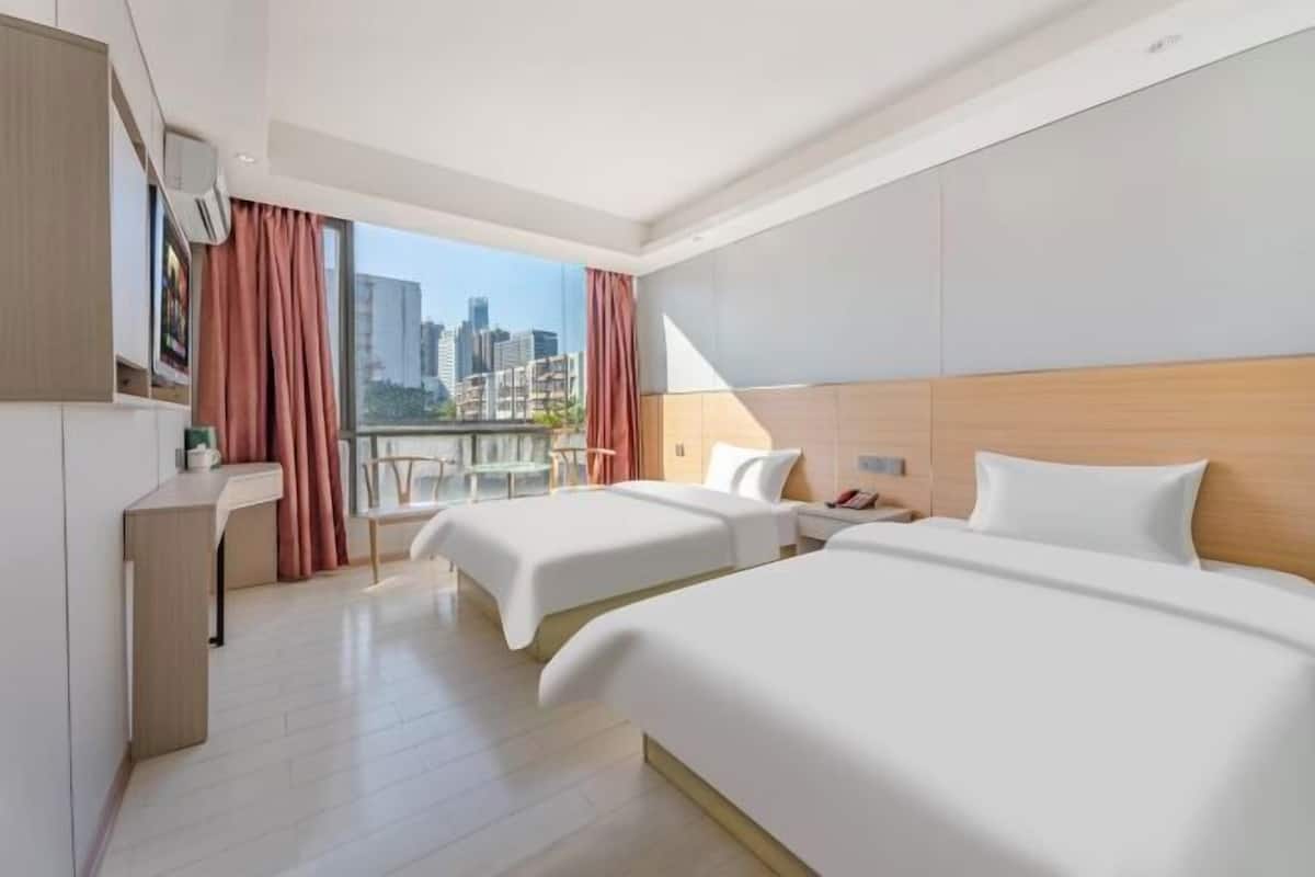 A hotel room with two beds, a desk, a chair, a window with curtains, and a city view.