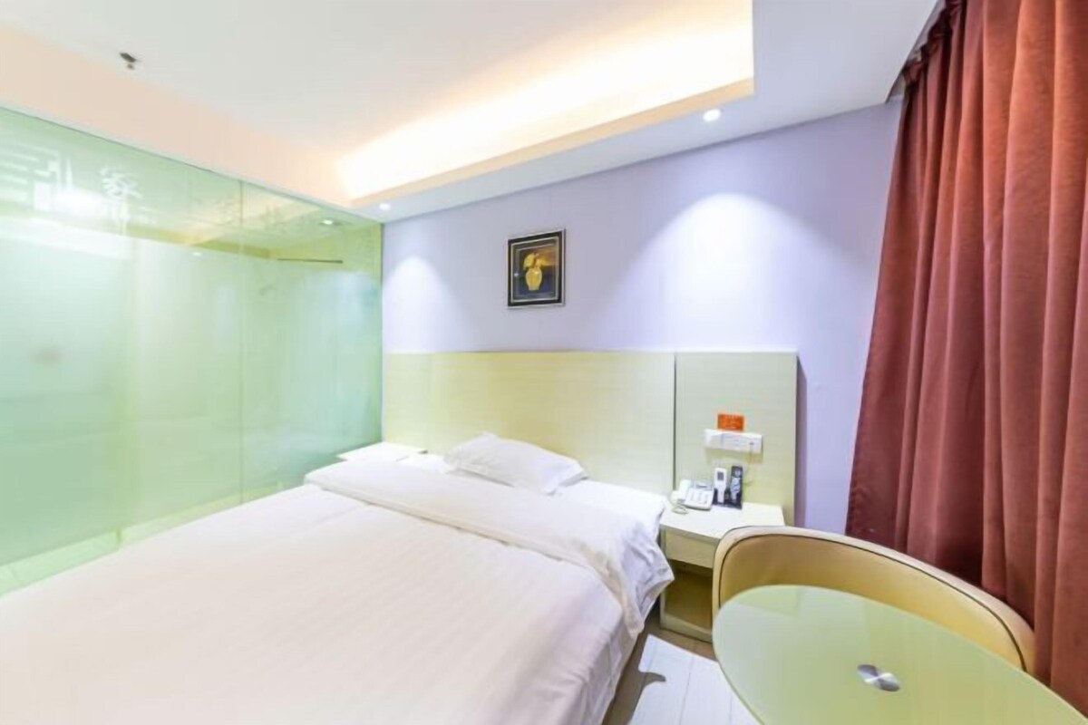 A modern hotel room with a large bed, a glass partition, a bedside table with a lamp, and a mirror.