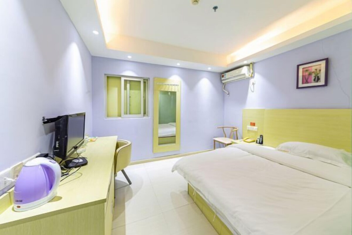 A hotel room with a bed, a desk with a computer and a kettle, a mirror, and a window.