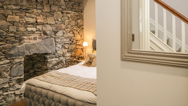 A bedroom with a stone fireplace, a bed with a plaid bedspread, and a white wall with a mirror and a wooden staircase. - The Haven (Windermere)