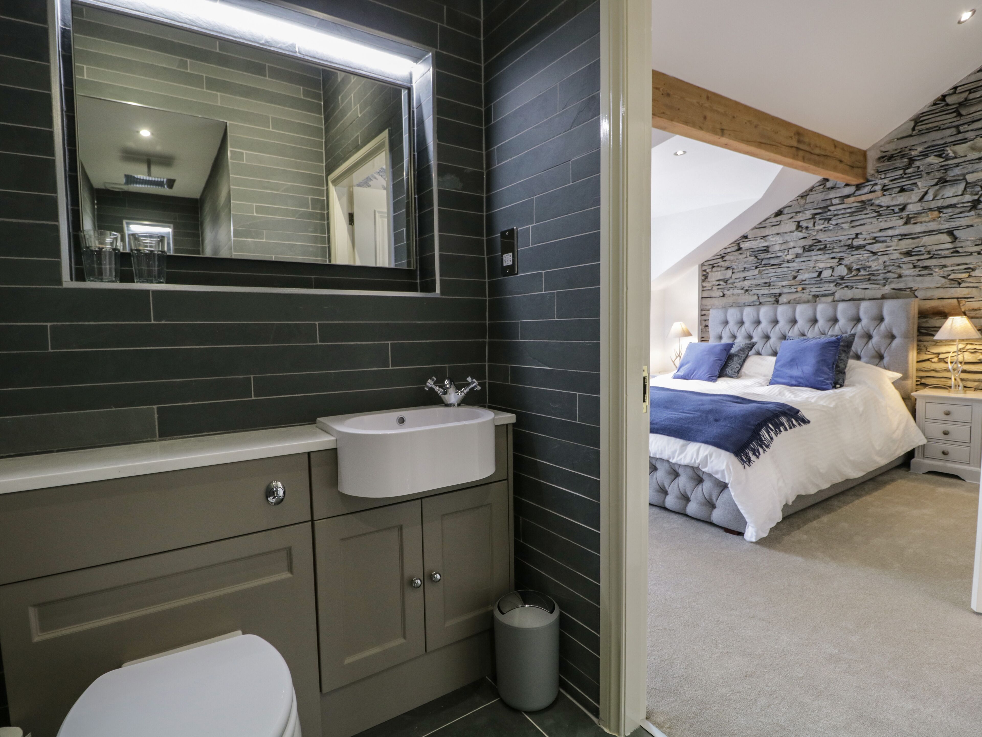 A modern bathroom with a large mirror, a white sink, and a toilet. The room features dark tiles and a stone wall.