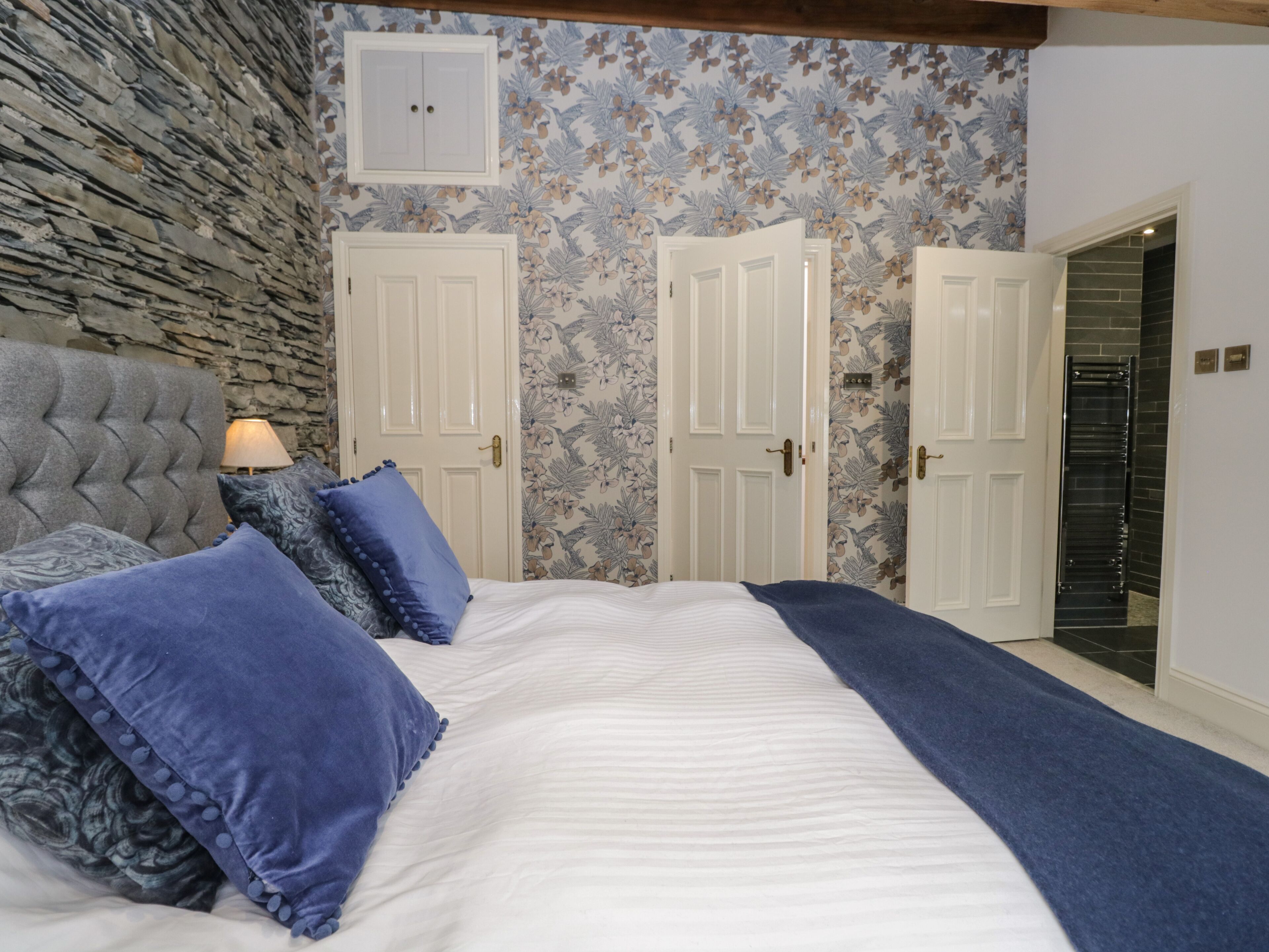 A bedroom with a stone wall, a bed with blue bedding, two white doors, and a small cabinet above one door.