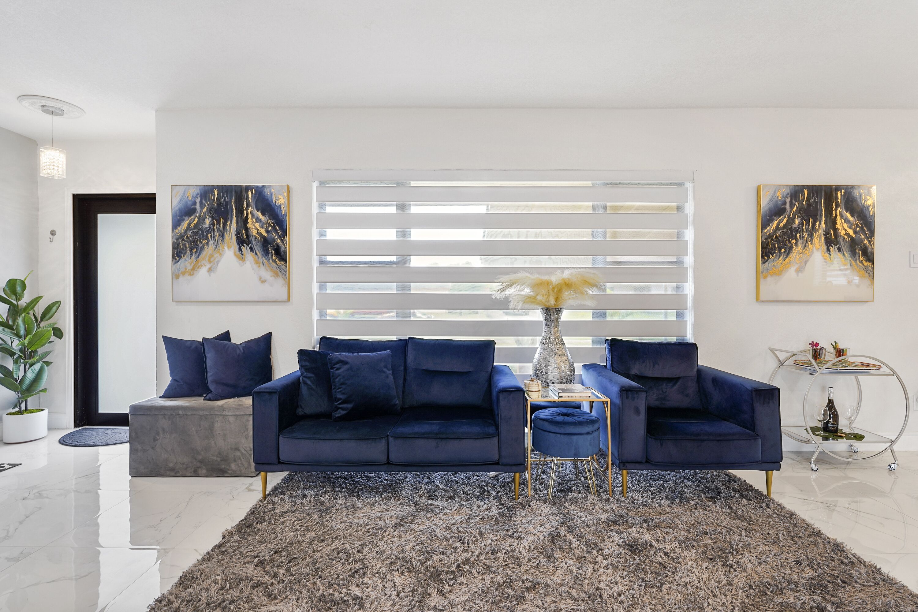 Una sala de estar moderna con un sofá azul oscuro, un reposapiés redondo y una mesa de centro de vidrio. En las paredes, dos cuadros abstractos, una planta en maceta y una mesita auxiliar metálica con objetos decorativos.