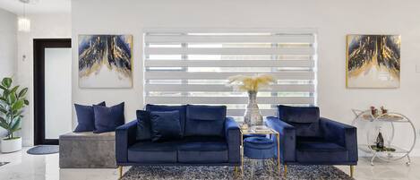 Una sala de estar moderna con un sofá azul oscuro, un reposapiés redondo y una mesa de centro de vidrio. En las paredes, dos cuadros abstractos, una planta en maceta y una mesita auxiliar metálica con objetos decorativos.