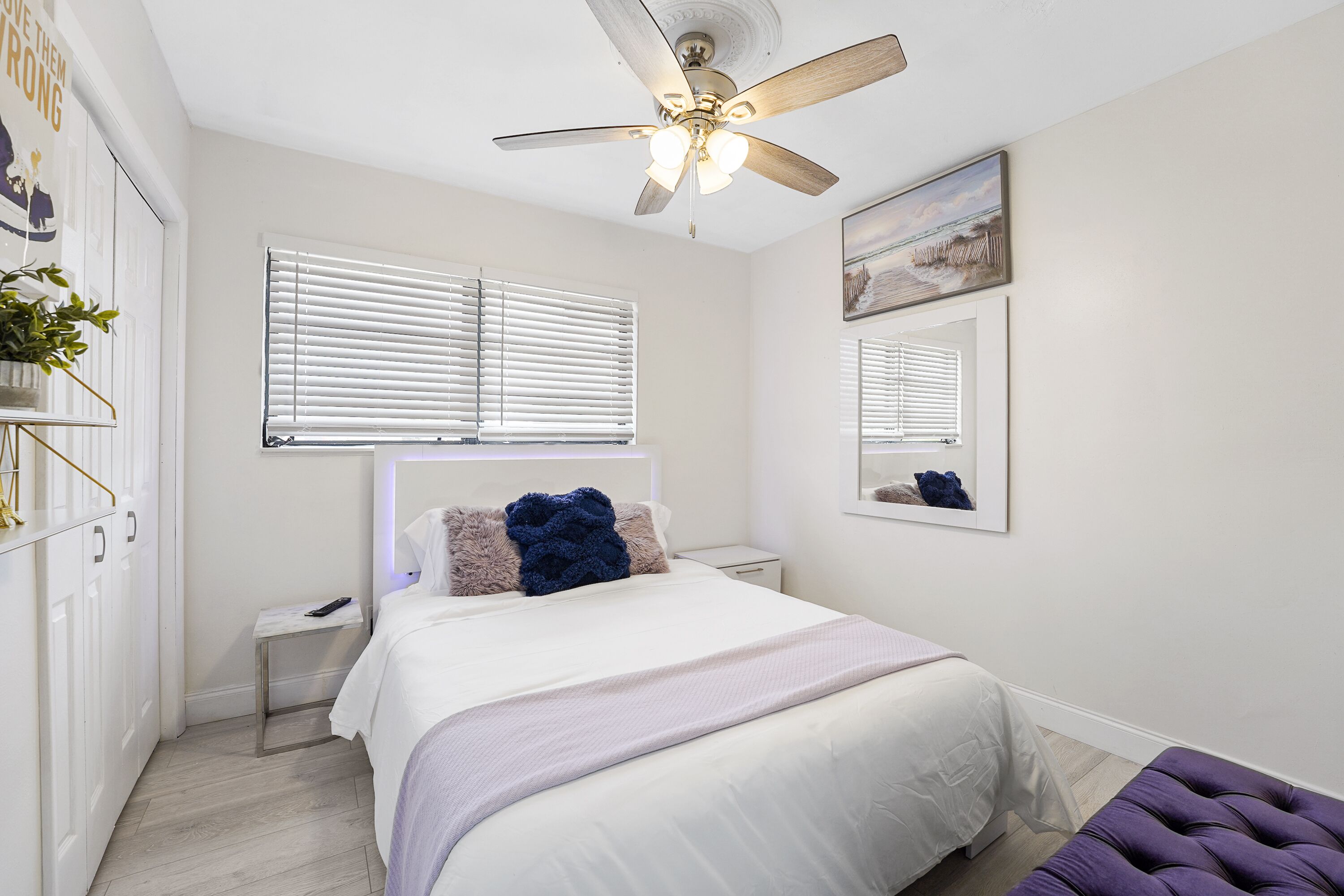 Un dormitorio con cama, ventilador de techo, mesita de noche, ventana con persianas y un taburete morado.
